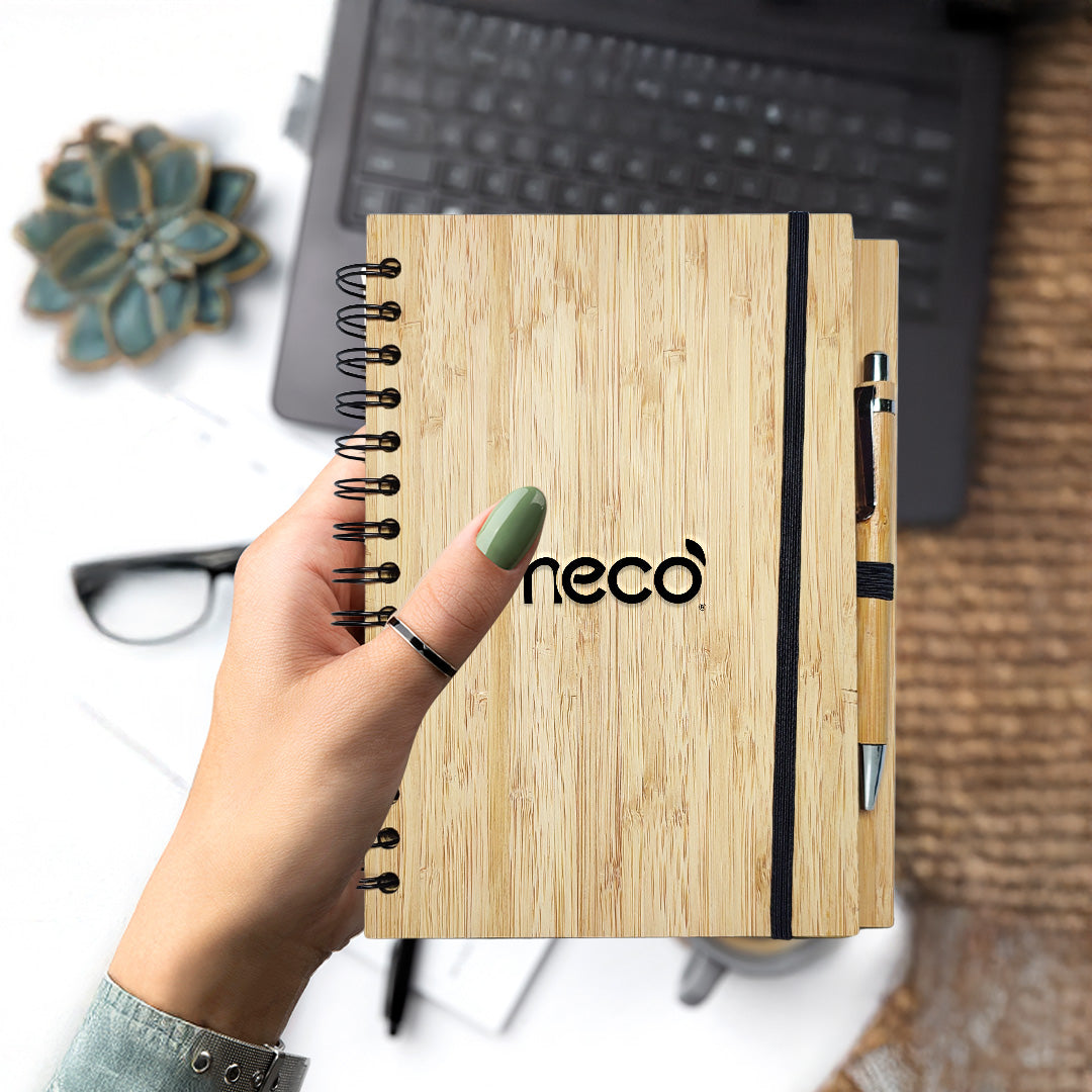 a hand is holding imeco bamboo diary with a pen against a blurred desk 