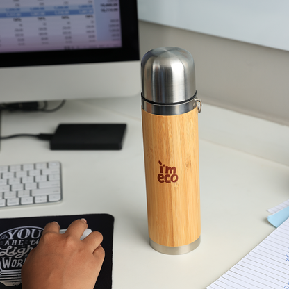 A bamboo thermos flask is placed on a work desk beside a desktop and a notebook.