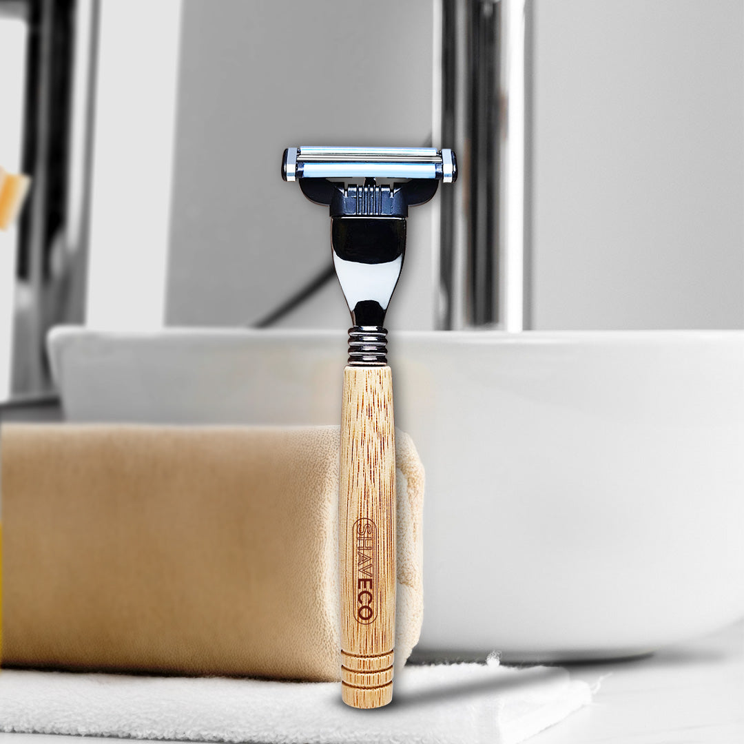 A bamboo razor is placed beside a towel and bathroom basin. It is engraved with the tag "SHAVECO".