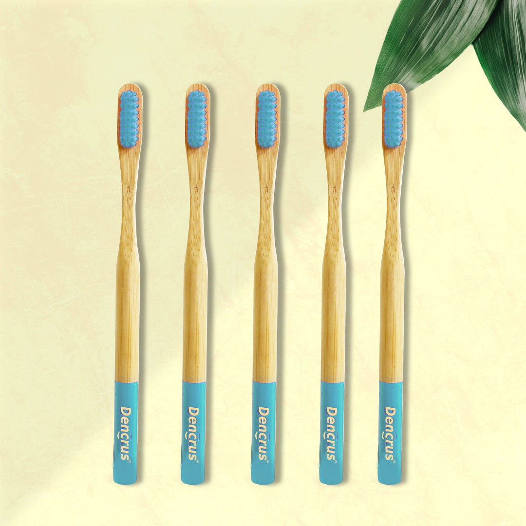 Five ORBY Plus bamboo toothbrushes with blue bristles and bamboo handles, displayed on a beige background with a green leaf to the side.